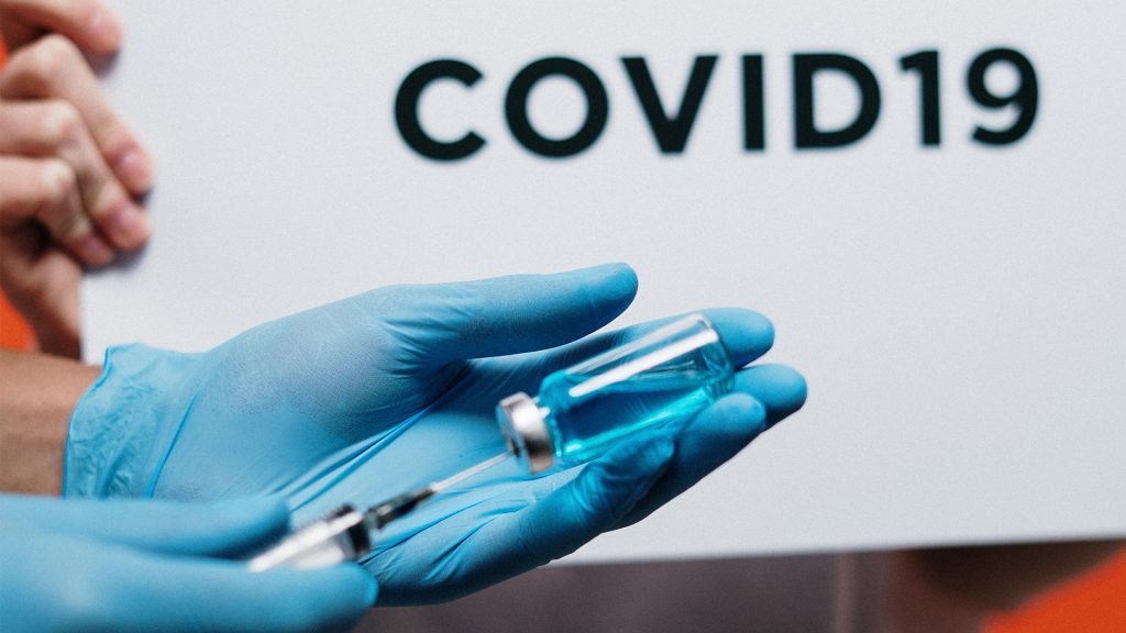 Impfspritze wird von Händen in Handschuhen gehalten, dahinter der Schriftzug COVID19