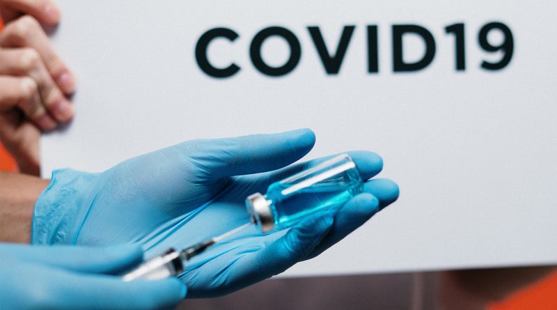 Impfspritze wird von Händen in Handschuhen gehalten, dahinter der Schriftzug COVID19