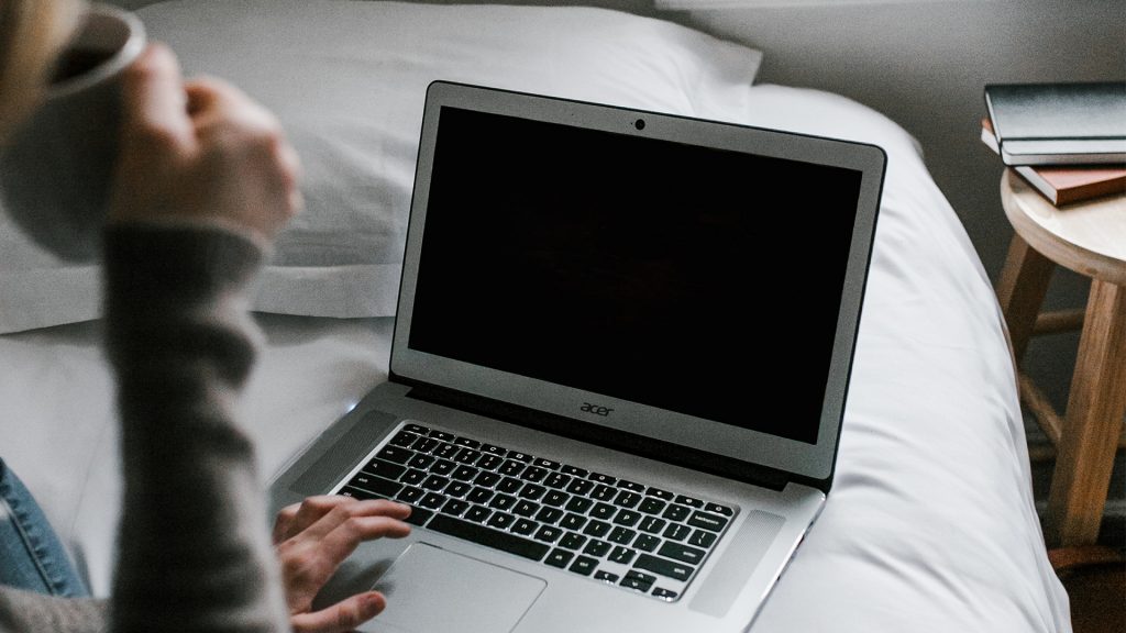 Frau trinkt aus einer Tasse auf dem Bett sitzend, davor ein Laptop, Bildschirm schwarz