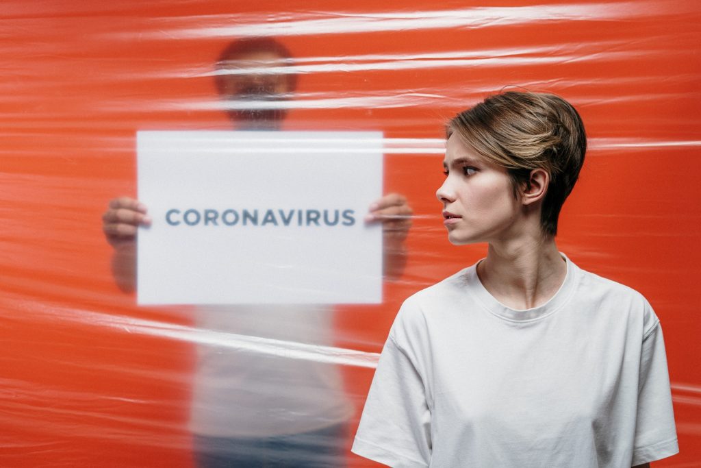 Mann steht mit Schild "Coronavirus" hinter einer durchsichtigen Barriere, davor steht eine Frau