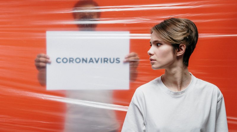 Mann steht mit Schild "Coronavirus" hinter einer durchsichtigen Barriere, davor steht eine Frau