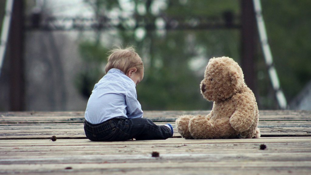 Ein kleines Kind sitzt gegenüber von einem Teddybär auf einer Holzterrasse