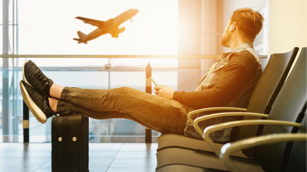 Mann sitzt am Flughafen, Füße auf dem Koffer, fliegendes Flugzeug im Hintergrund