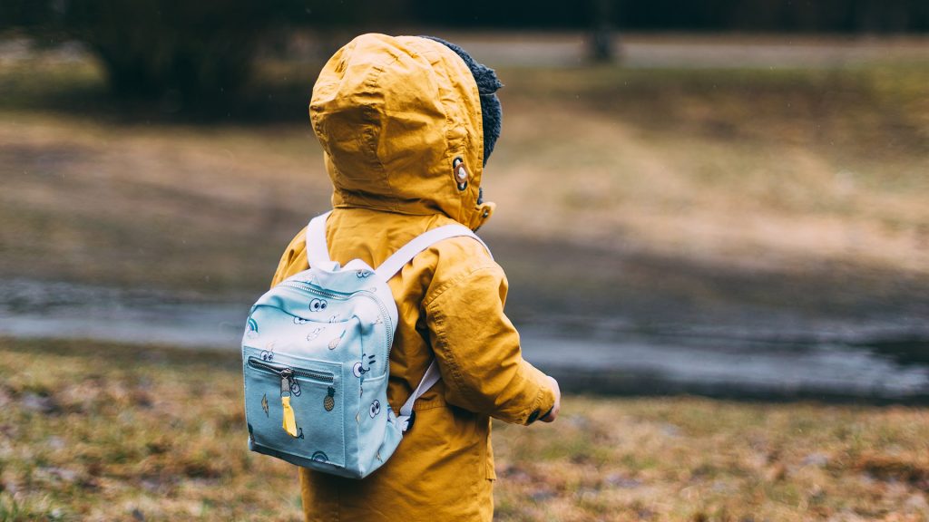 Kind mit gelber Regenjacke und rosa Schulrucksack, von hinten fotografiert