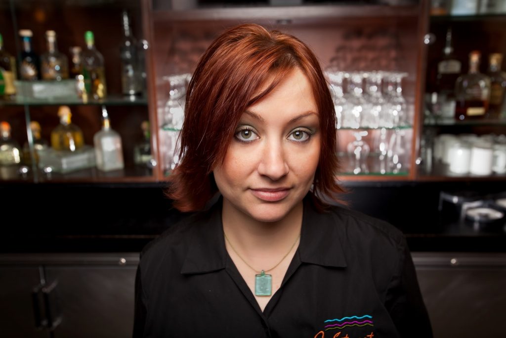 Frau mit schulterlangen Haaren und schwarzem Oberteil steht vor einer Bar
