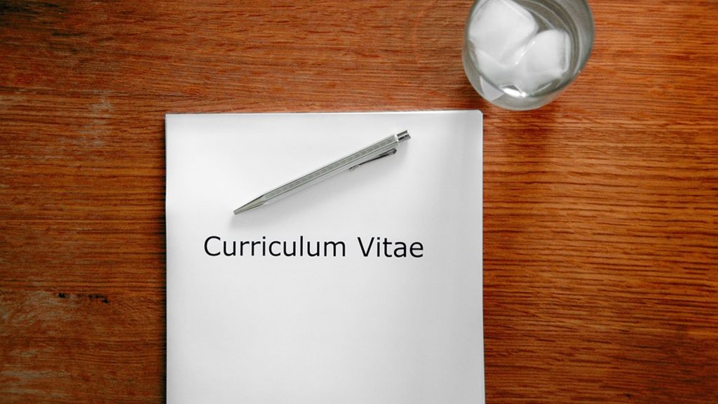 Zettel mit Aufschrift "Curriculum Vitae" liegt auf einem Holztisch, daneben ein Glas mit Eiswürfeln