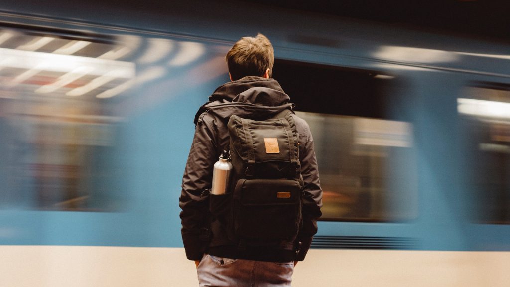 Mensch mit Rucksack steht am Bahnsteig, ein Zug fährt schnell vorbei