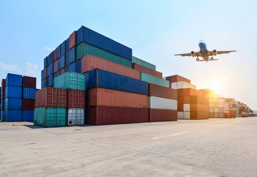 Containerstapel am Hafen, darüber fliegt ein FLugzeug