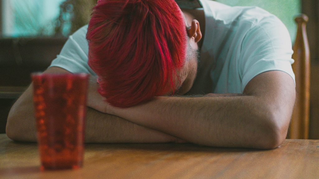 Mann mit weißem Shirt hat Arme auf dem Tisch verschränkt und Kopf in die Arme gelegt
