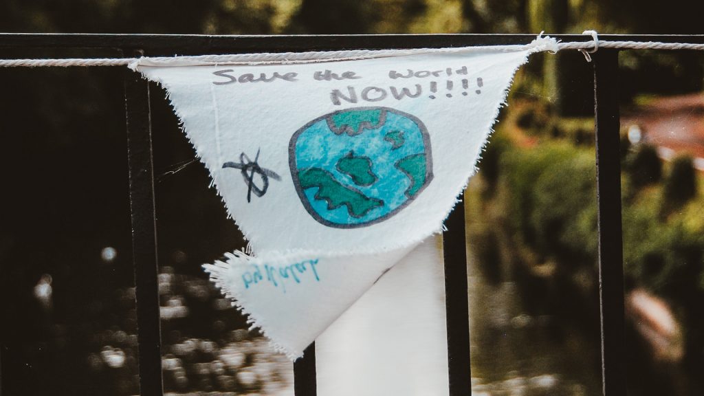 Weißes Stück Stoff mit Aufschrift "Save the World NOW!!!" hängt an einem Brückengeländer