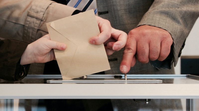 Drei Hände mit einem Kuvert in der Hand, das in eine Wahlurne geworfen wird