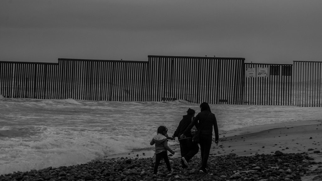 Drei Menschen an der Grenzen zwischen den USA und Mexiko am Meer, im Hintergrund der Grenzzaun