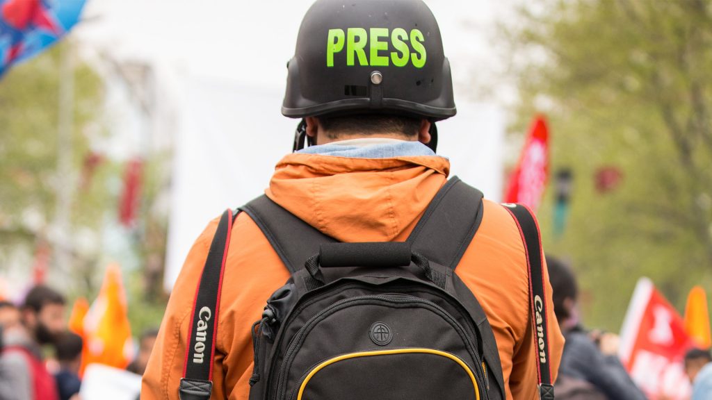 Journalist mit orangener Jacke und schwarzem Rucksack und Helm mit Aufschrift PRESS von hinten fotografiert
