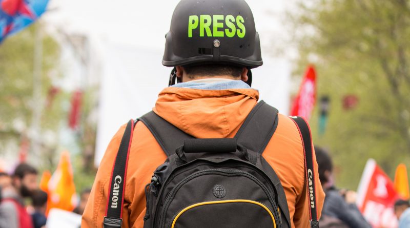 Journalist mit orangener Jacke und schwarzem Rucksack und Helm mit Aufschrift PRESS von hinten fotografiert