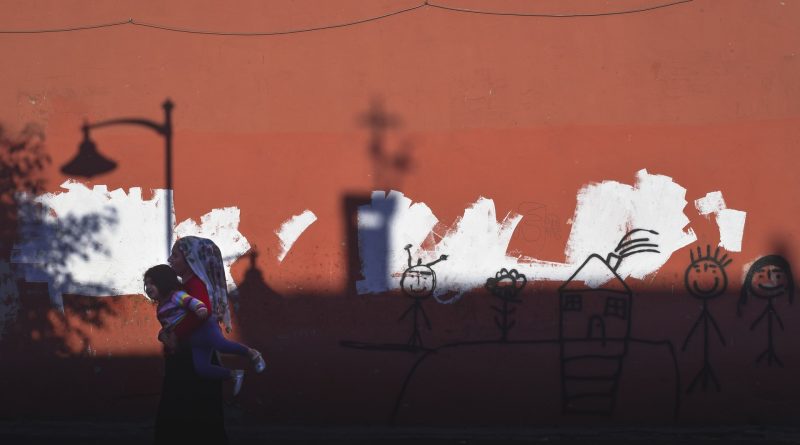 Frau mit Kind auf dem Arm flieht, im Hintergrund sieht man eine rote Mauer