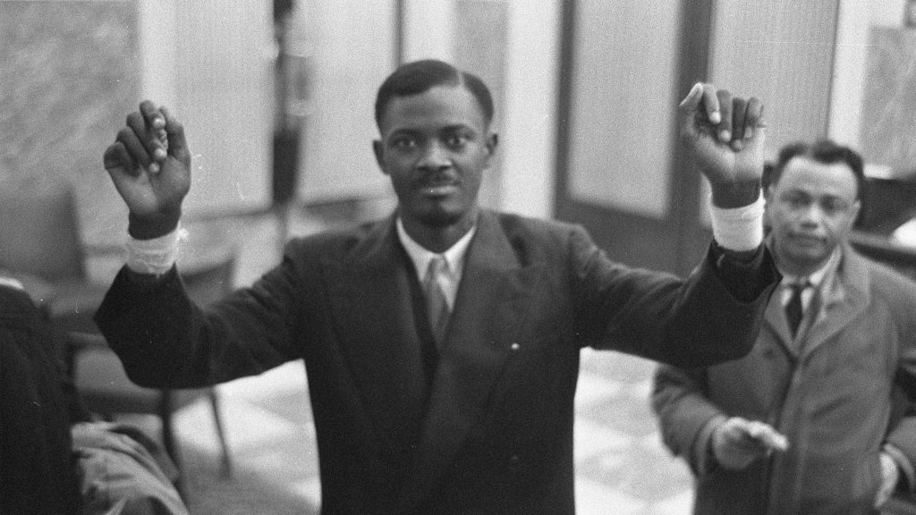 Patrice Lumumba im Jahr 1960 im Anzug, beide Hände angewinkelt in die Luft gestreckt
