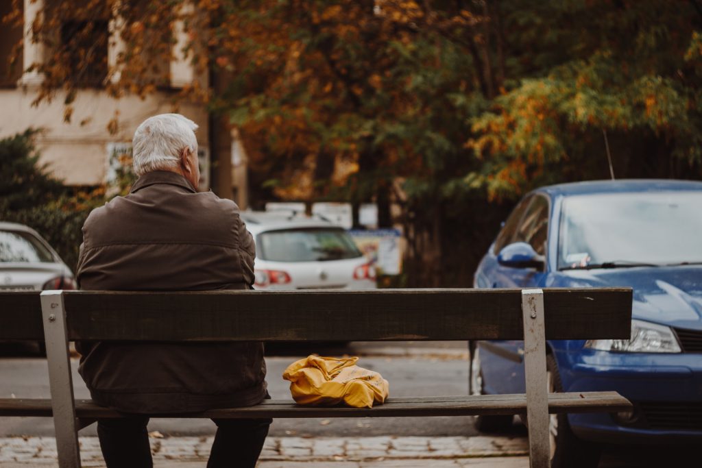 Mann mit grauen Haaren sitzt auf einer Bank vor einem Parkplatz