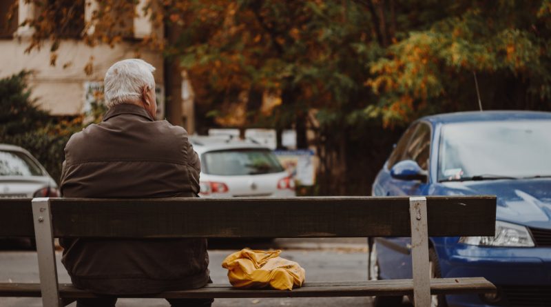 Mann mit grauen Haaren sitzt auf einer Bank vor einem Parkplatz