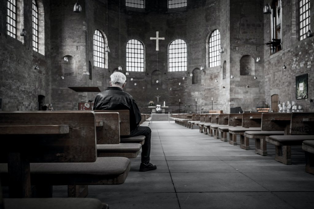 Ein Mann mit weißen Haaren sitzt alleine in einer großen Kirche, Blick nach vorne auf dem Alter