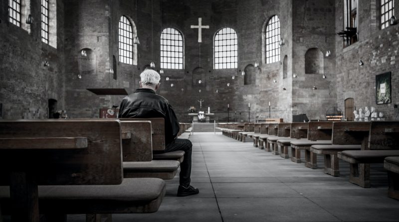 Ein Mann mit weißen Haaren sitzt alleine in einer großen Kirche, Blick nach vorne auf dem Alter