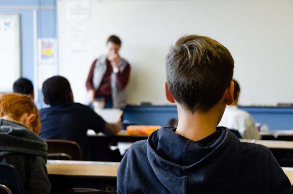 Kind mit kurzen Haaren auf einer Schulbank, im unscharfem Hintergrund steht eine Lehrperson