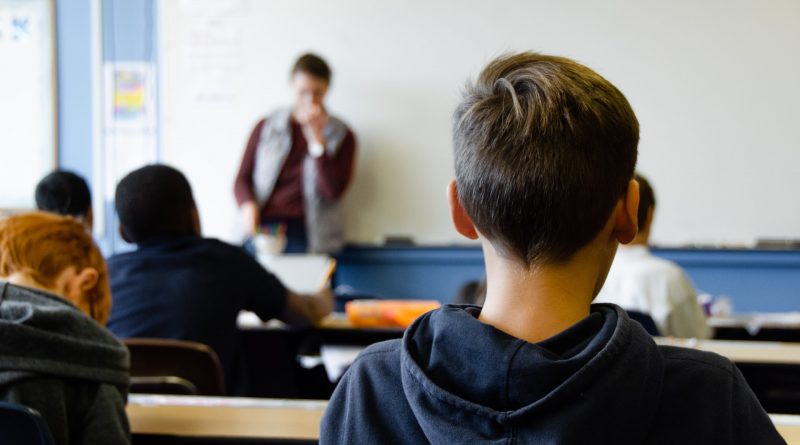 Kind mit kurzen Haaren auf einer Schulbank, im unscharfem Hintergrund steht eine Lehrperson
