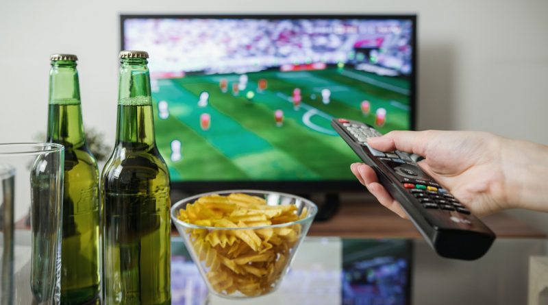 Person hält Fernbedienung, im Hintergrund läuft Fußball im Fernsehen. Auf dem Tisch davor stehen Bier und Snacks