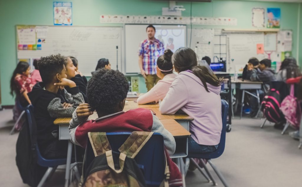 Klassenzimmer mit Schüler*innen an Tischen sitzend und einem Lehrer, der steht