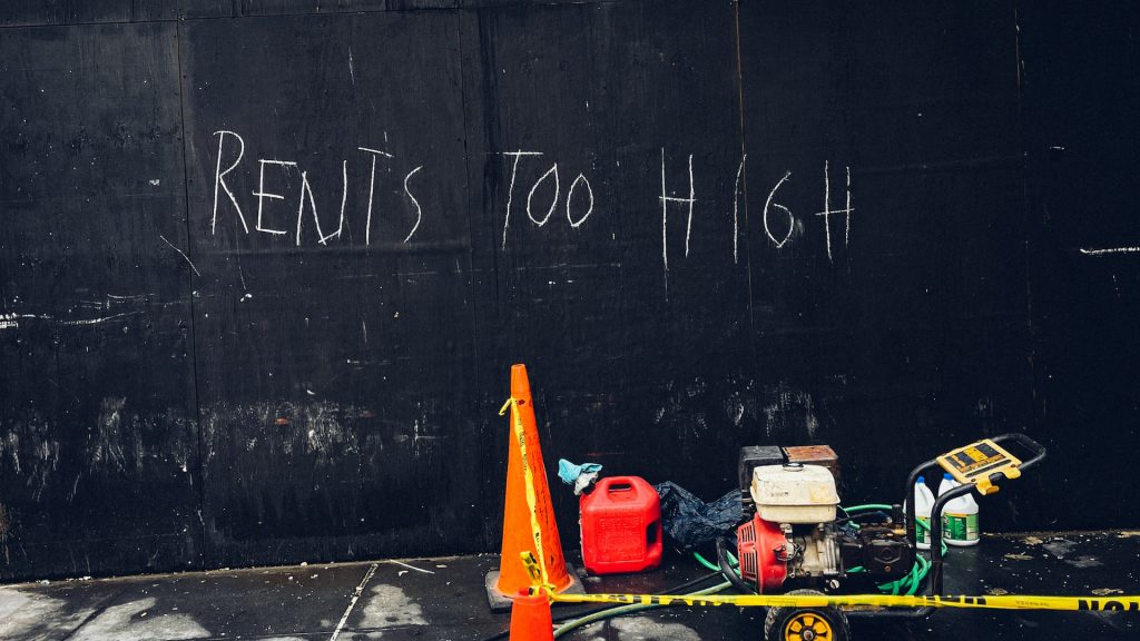 Schwarze Wand mit weißer Aufschrift "Rents too high", Davor liegen Baustellen-Geräte