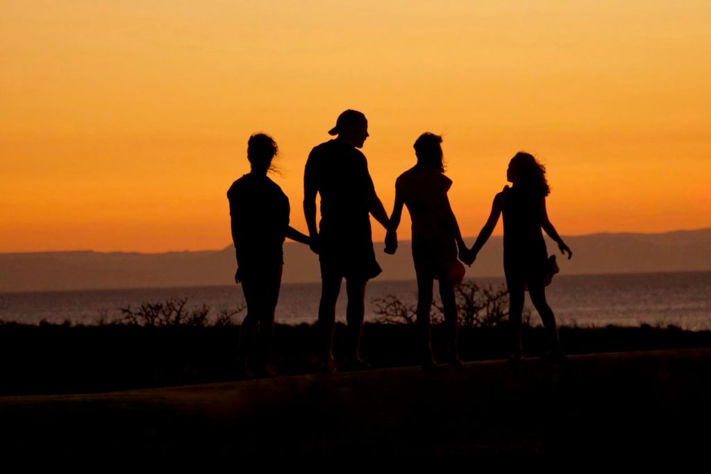 Vier Personen als Silhouetten vor einem Sonnenuntergang halten sich an den Händen