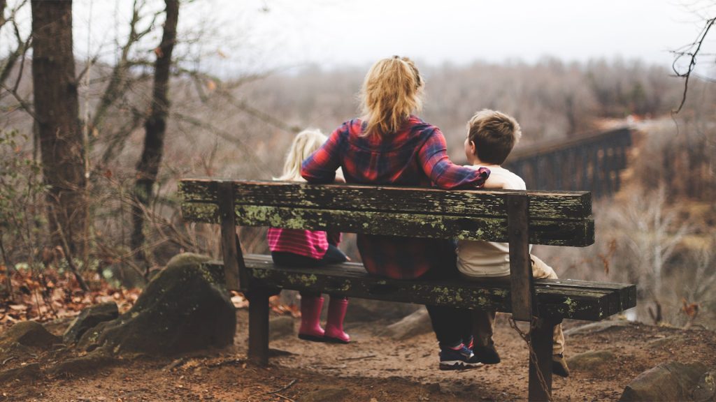 Frau sitzt mit zwei Kindern auf einer Bank im Wald.