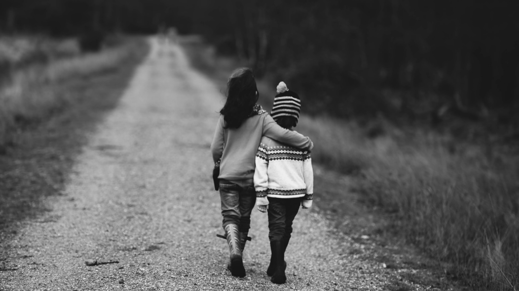 Zwei Kinder gehen auf einem Weg, eines hat die Hand um das andere gelegt. Schwarz-weiß