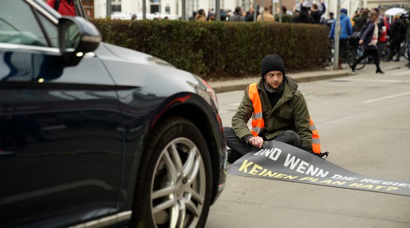 Schwarzes Auto steht vor einem Mitglied der Letzten Generation, das auf der Straße sitzt