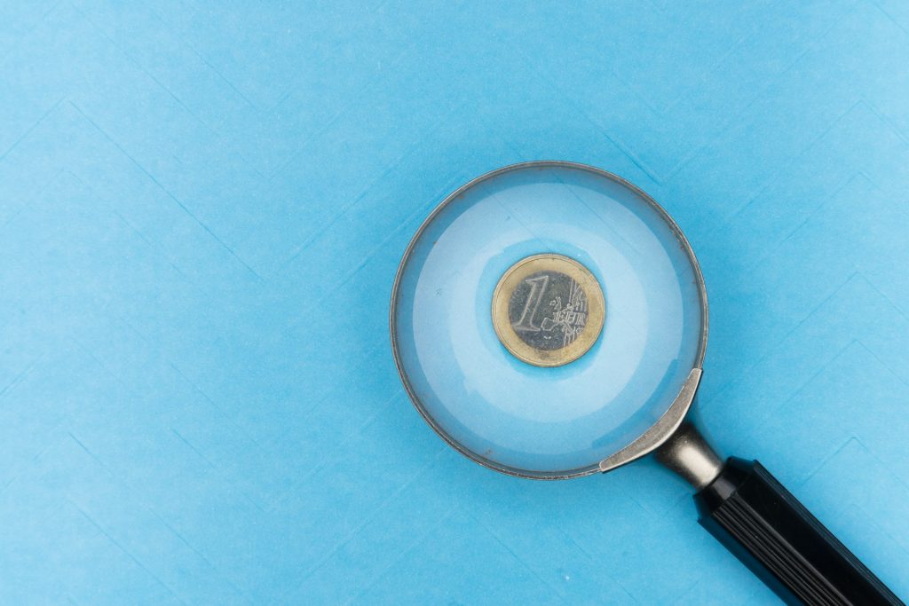 Ein 1-Euro-Stück, auf das mit einer Lupe geschaut wird, blauer Hintergrund