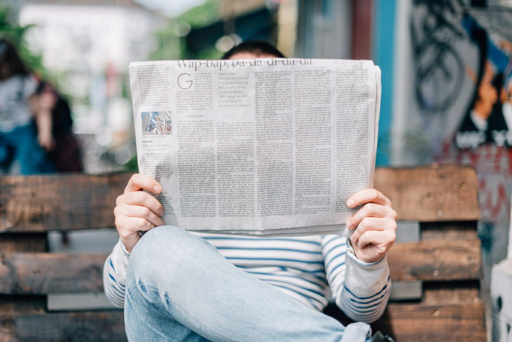 Person sitzt auf einer Holzbank und hält eine Zeitung in den Händen, die das Gesicht der Person verdeckt.