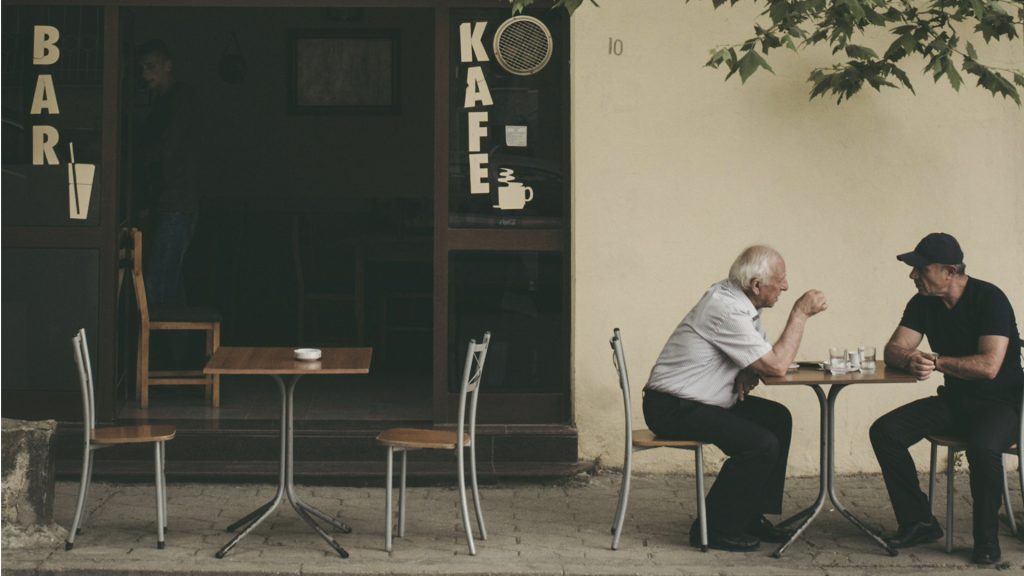 Zwei Männer sitzen an einem kleinen Tisch im Außenbereichs eines Cafés unter unterhalten sich.