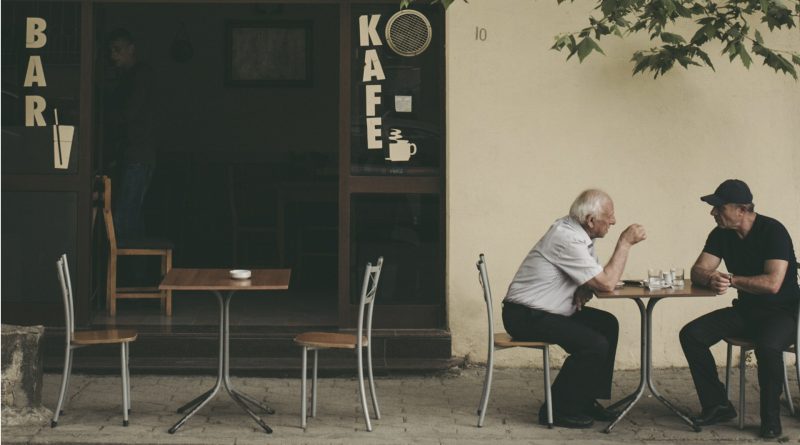 Zwei Männer sitzen an einem kleinen Tisch im Außenbereichs eines Cafés unter unterhalten sich.