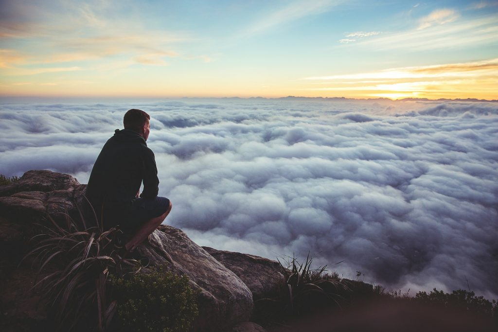 Person sitzt auf einem Felsen und schaut in die Ferne auf ein Wolkenmeer und den Sonnenuntergang